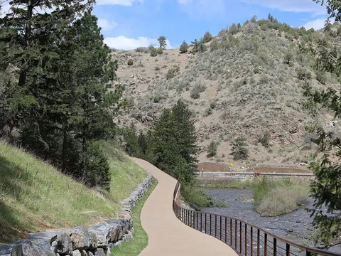 The paved Clear Creek Trail curving along a steel railing above the creek in Clear Creek Canyon, with US 6 across the water